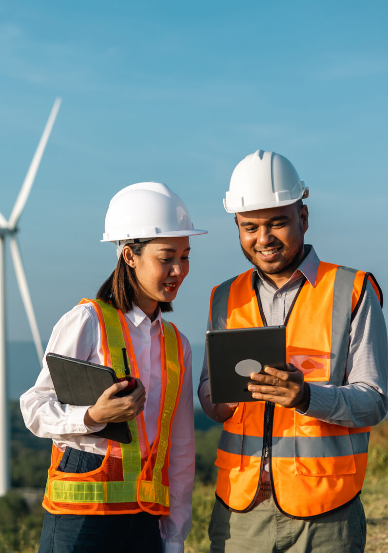 Two engineers of different ethnicities reviewing a tablet together in front of a wind farm. 