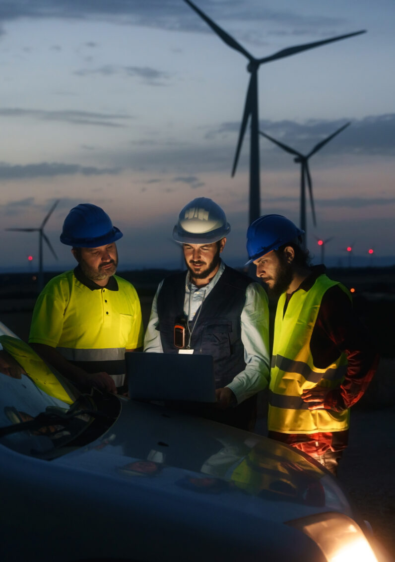 Three electrical sector employees discussing work in front of some wind turbines at night
