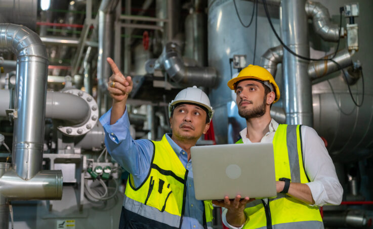 Deux ingénieurs électriciens professionnels en uniforme de sécurité travaillant ensemble dans la salle de contrôle d’un site industriel.